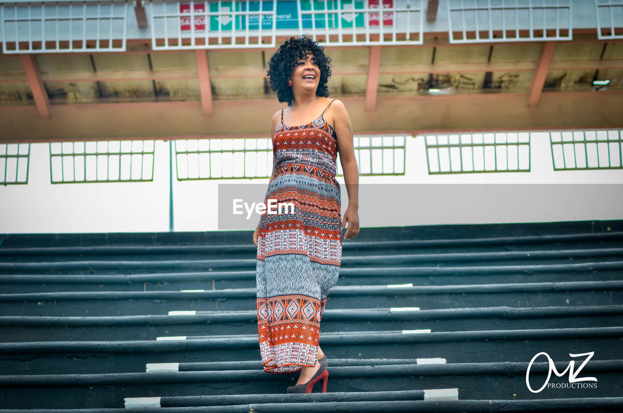 FULL LENGTH OF YOUNG WOMAN STANDING AGAINST STAIRCASE