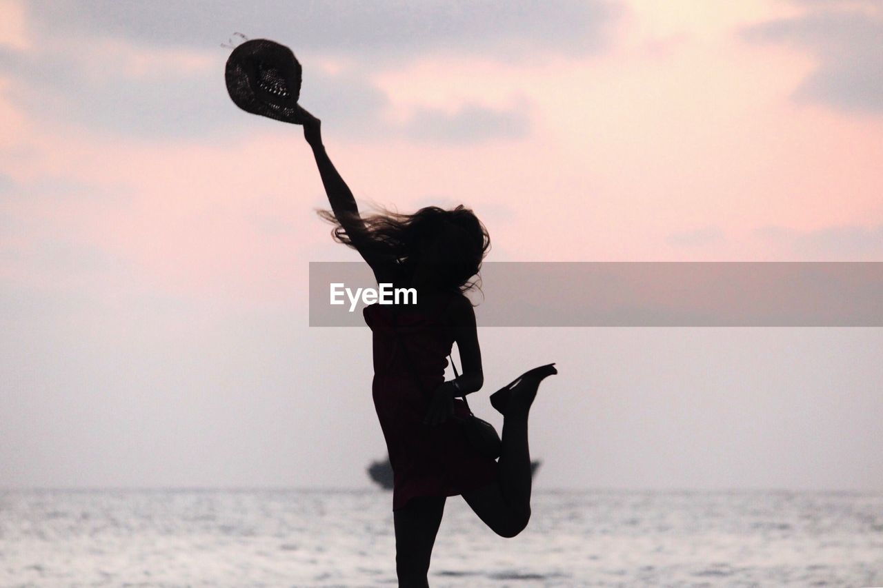 Silhouette woman holding hat while jumping at beach against sky during sunset