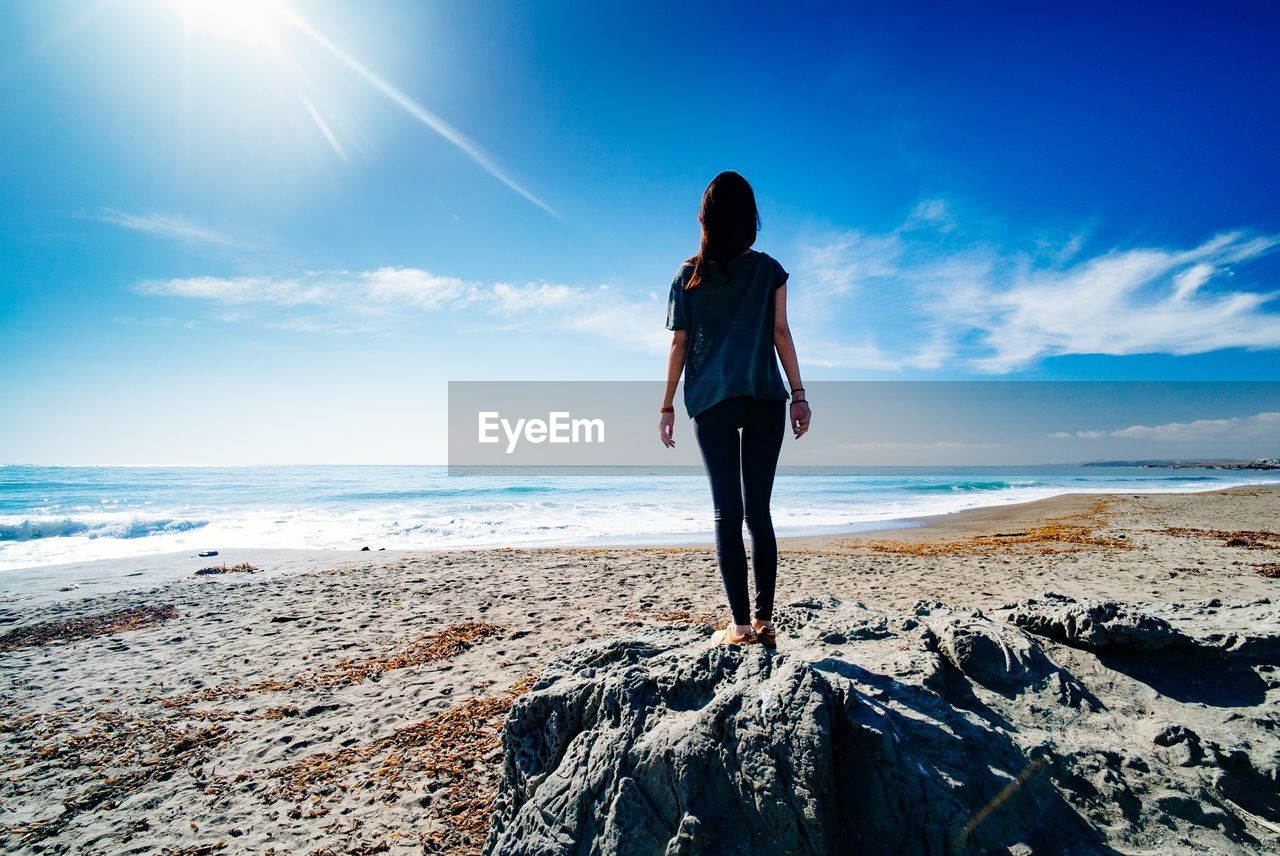 Rear view of woman standing on rock against blue sky at beach