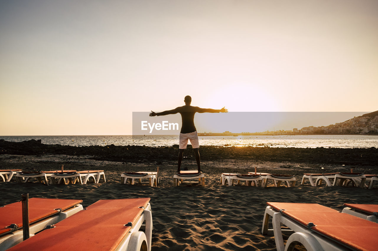 Rear view of silhouette young man with arms outstretched standing on deck chair at beach against clear sky during sunset