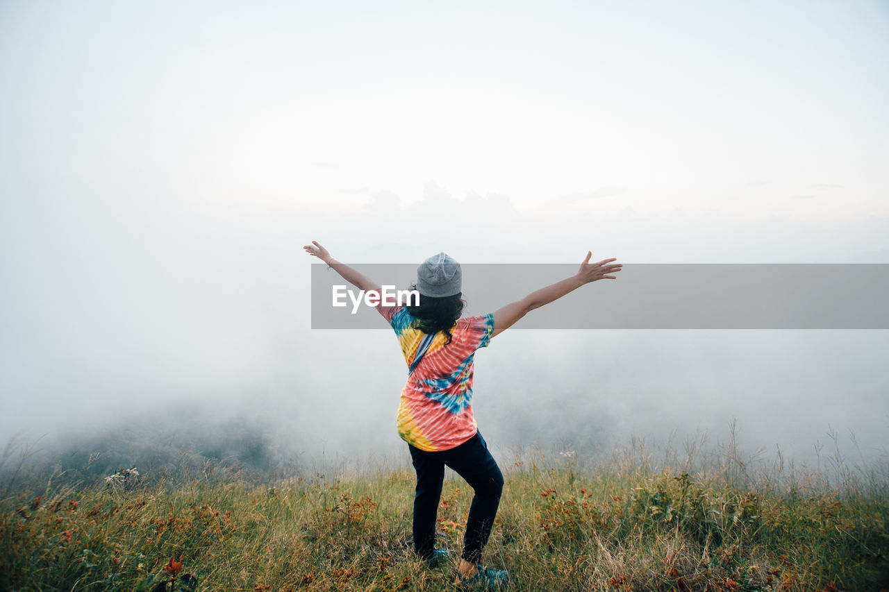 Rear view of woman with arms outstretched standing on mountain during foggy weather