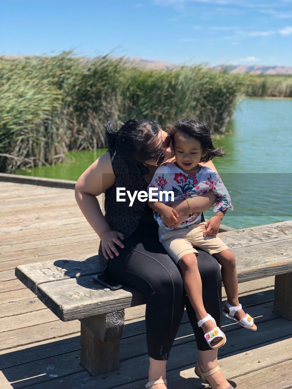 Mother kissing daughter while sitting on bench against lake