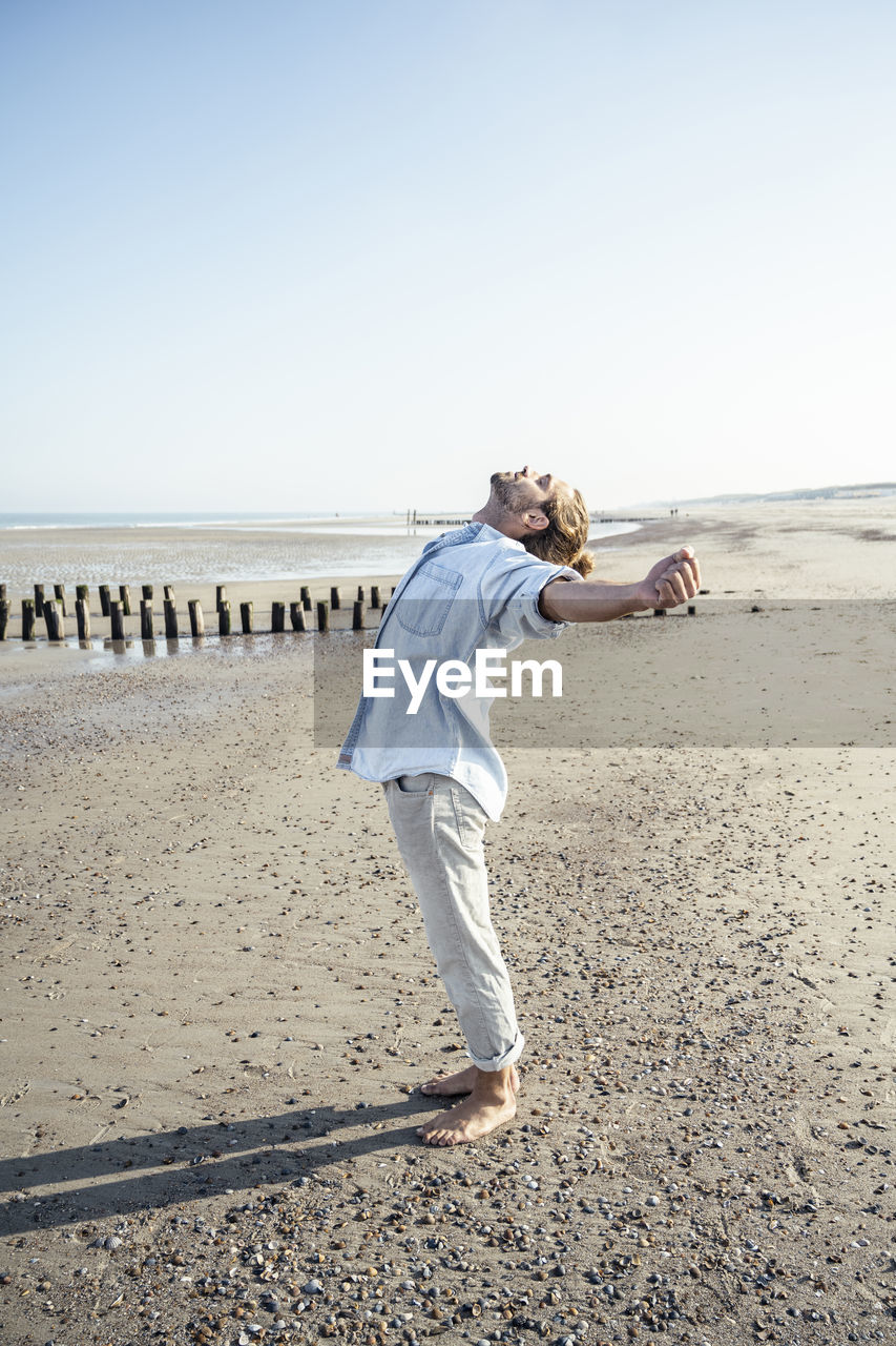 Young man with hands outstretched standing on beach during sunny day