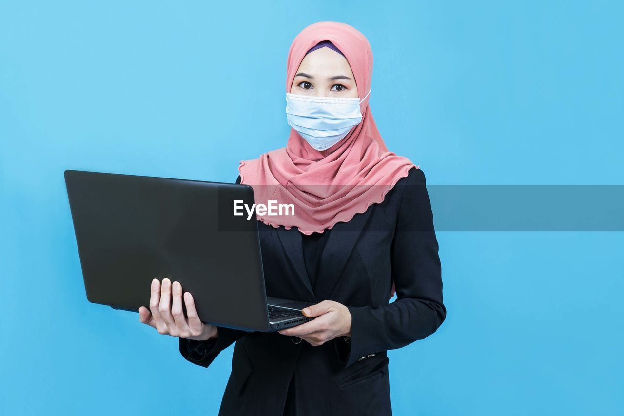 Muslim asian business woman with traditional dress wearing medical face mask  holding laptop