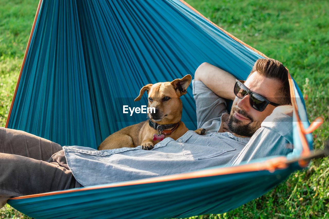Young handsome european man in sunglasses is resting in hammock with his cute little dog.