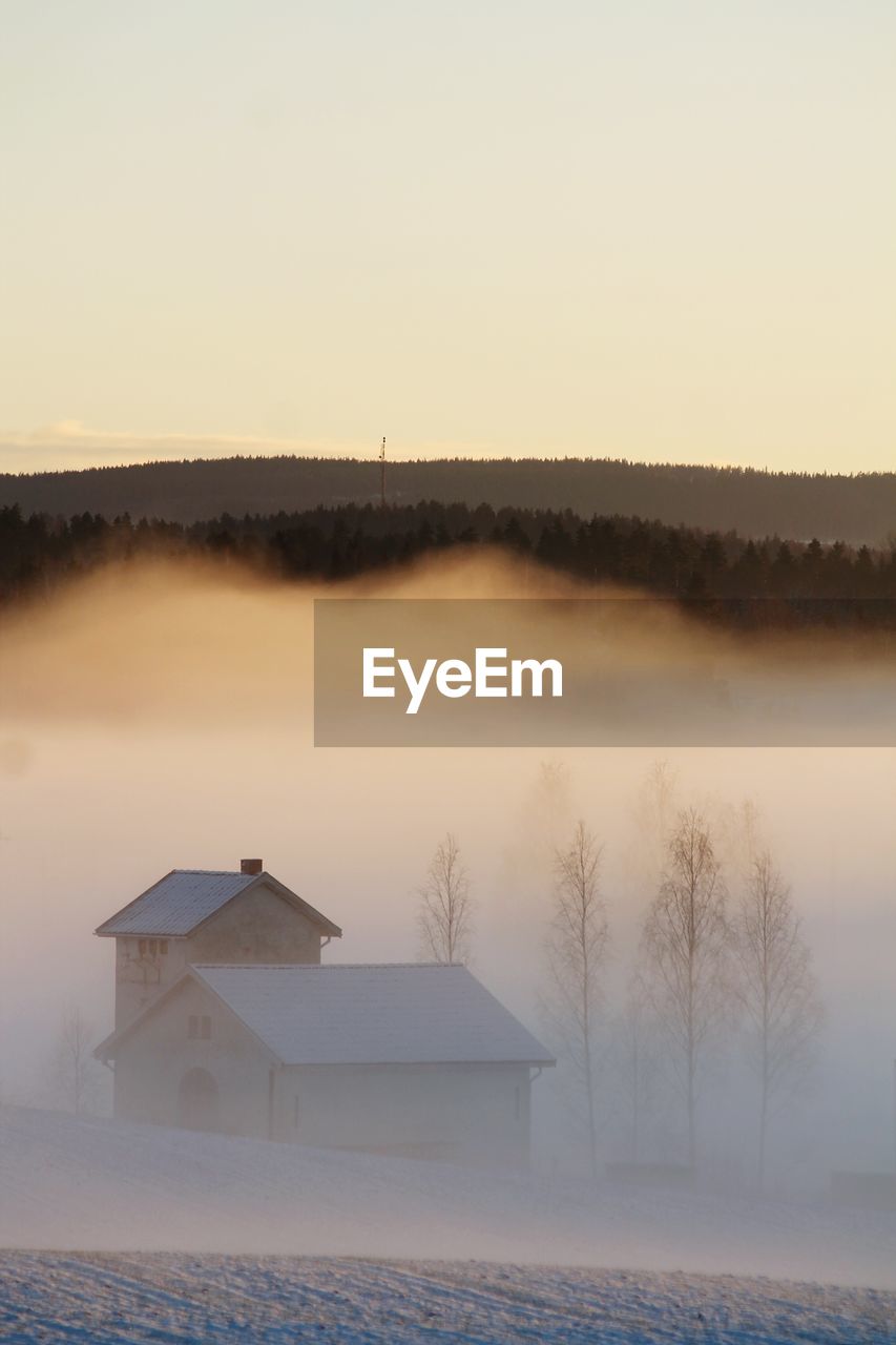 Houses amidst fog on snow covered field against sky during sunset