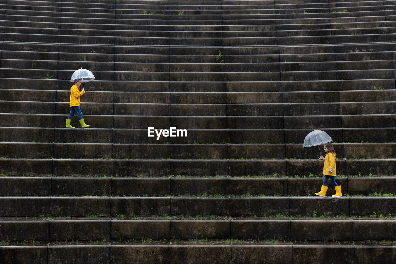 Side view of children in vivid yellow raincoats and rubber boots walking under umbrellas along wet stone stairs in street on rainy day