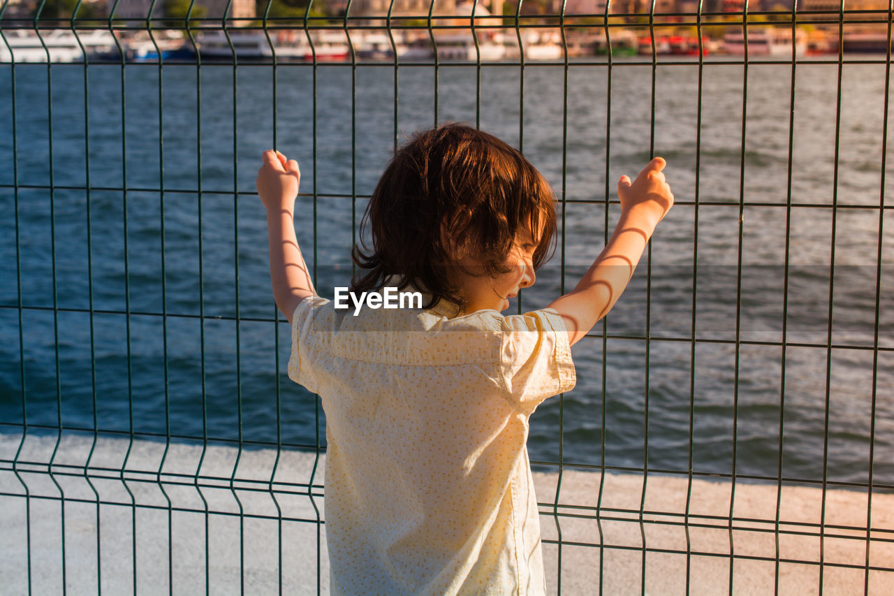 REAR VIEW OF BOY STANDING AGAINST FENCE