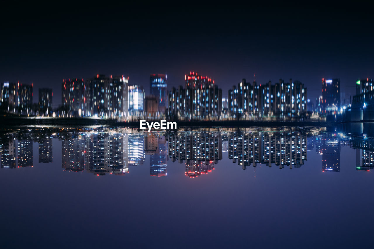 Illuminated modern buildings by sea against sky at night