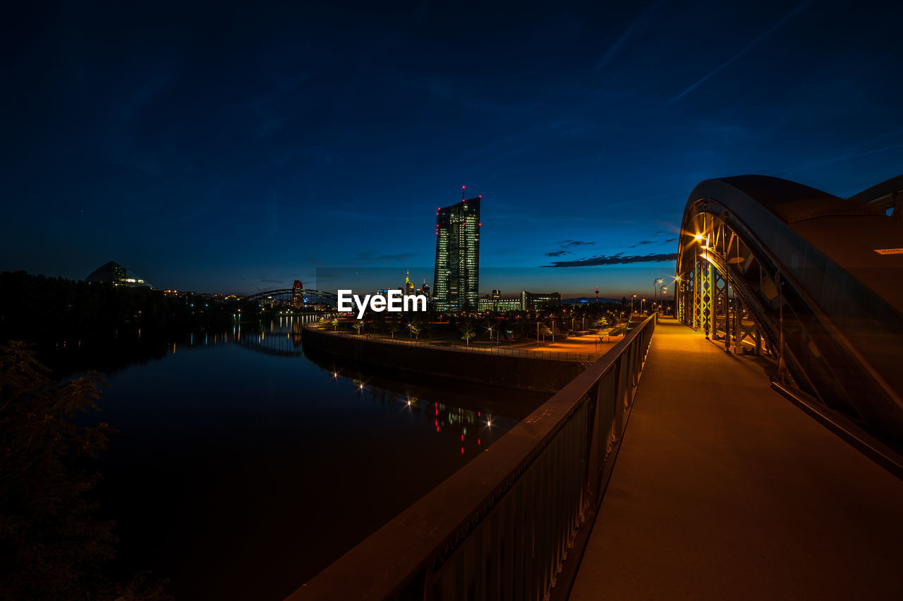 Illuminated bridge over river by buildings against sky at night