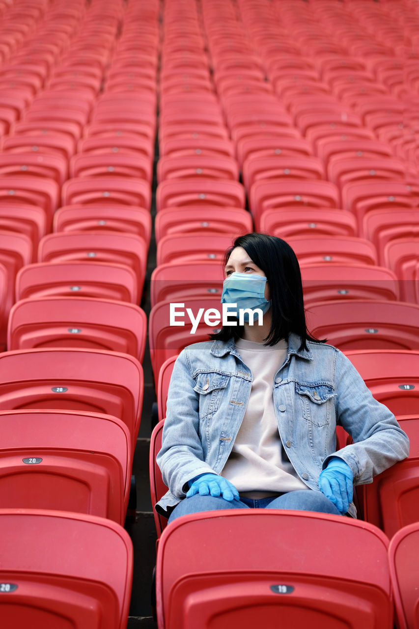 A female fan in a medical mask and rubber gloves sits alone in an empty stadium with red seats.