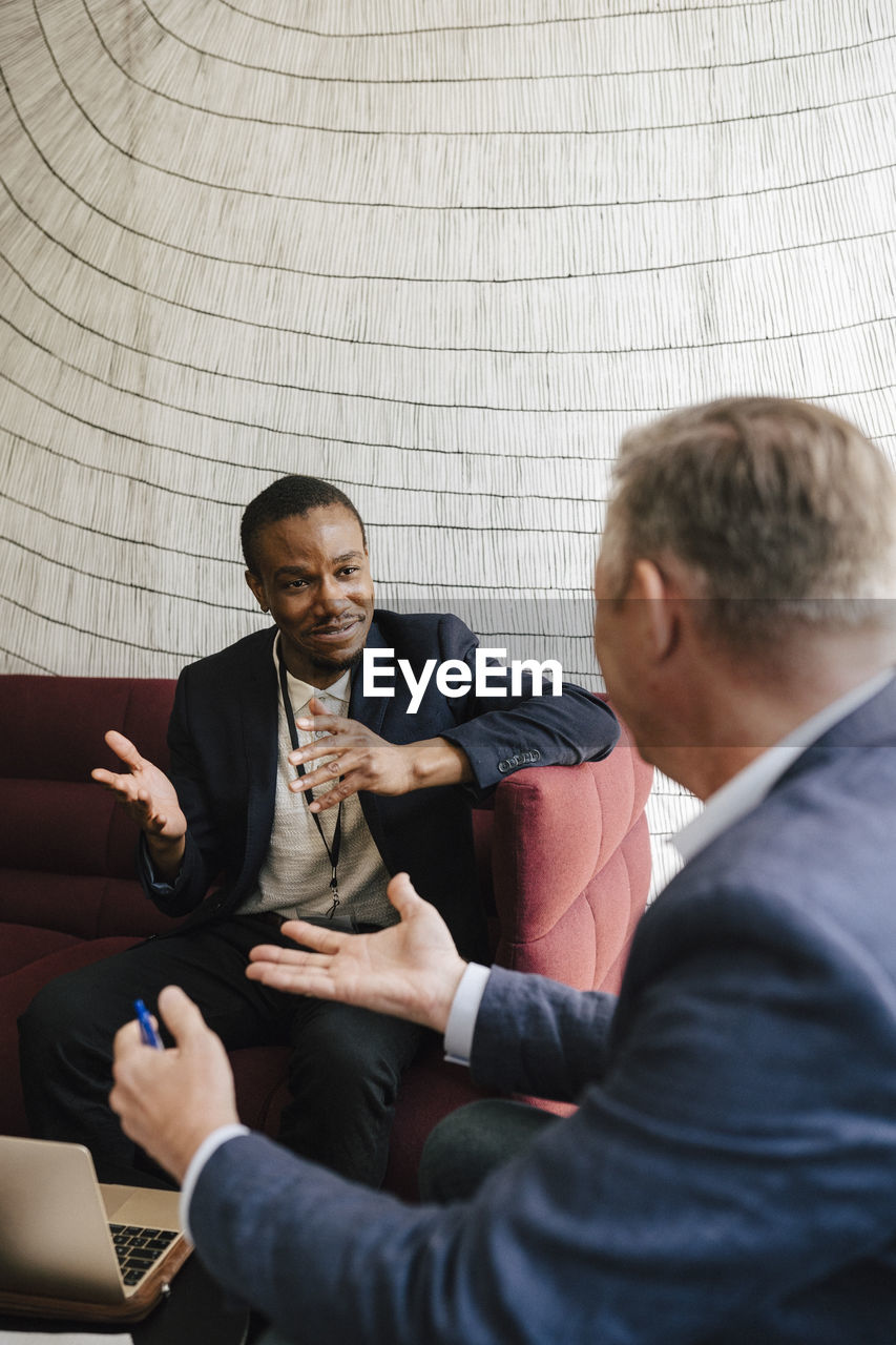 Male colleagues gesturing while discussing together sitting on sofa during networking event