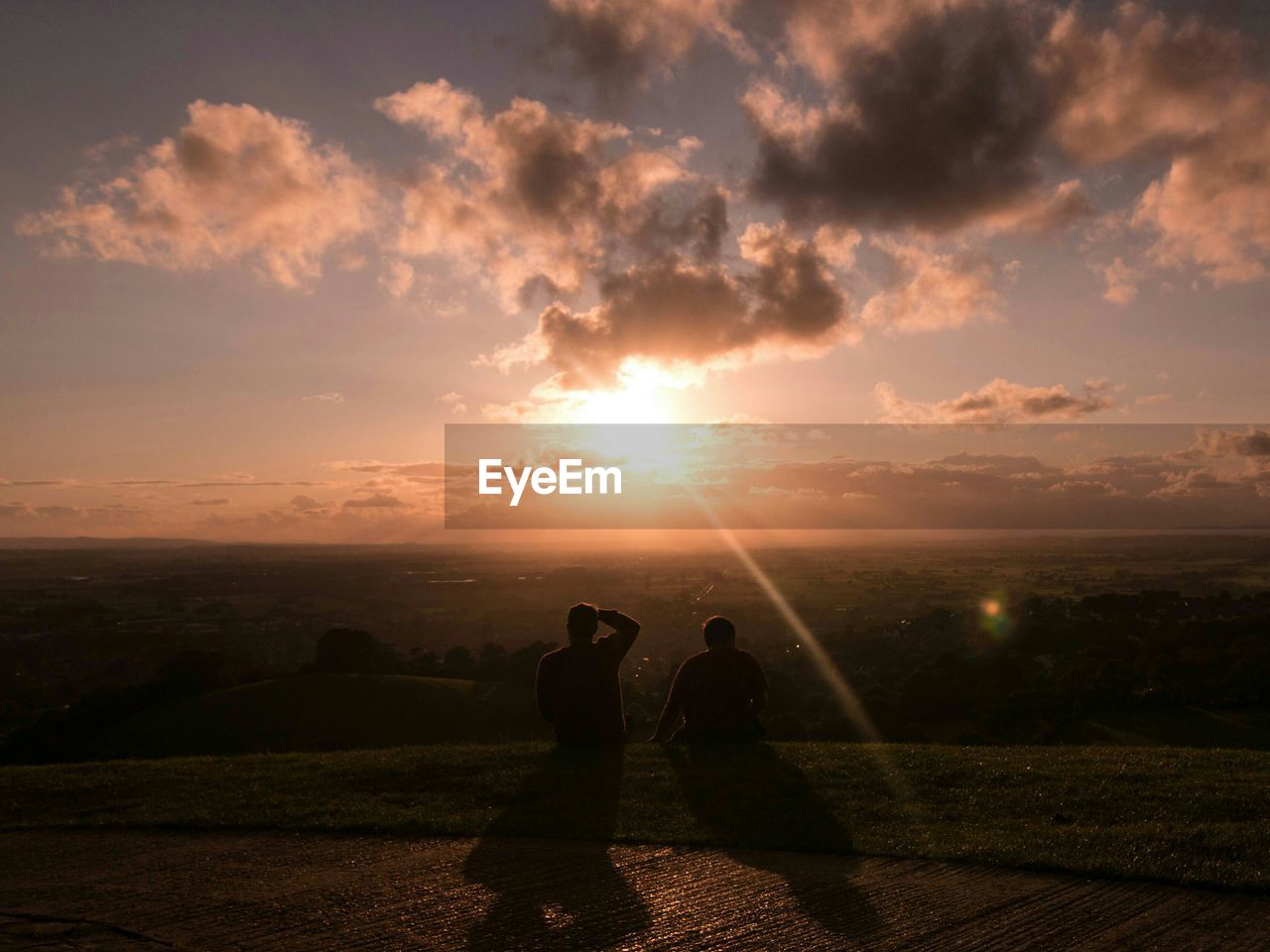 Rear view of men sitting on grassy field against sky during sunset