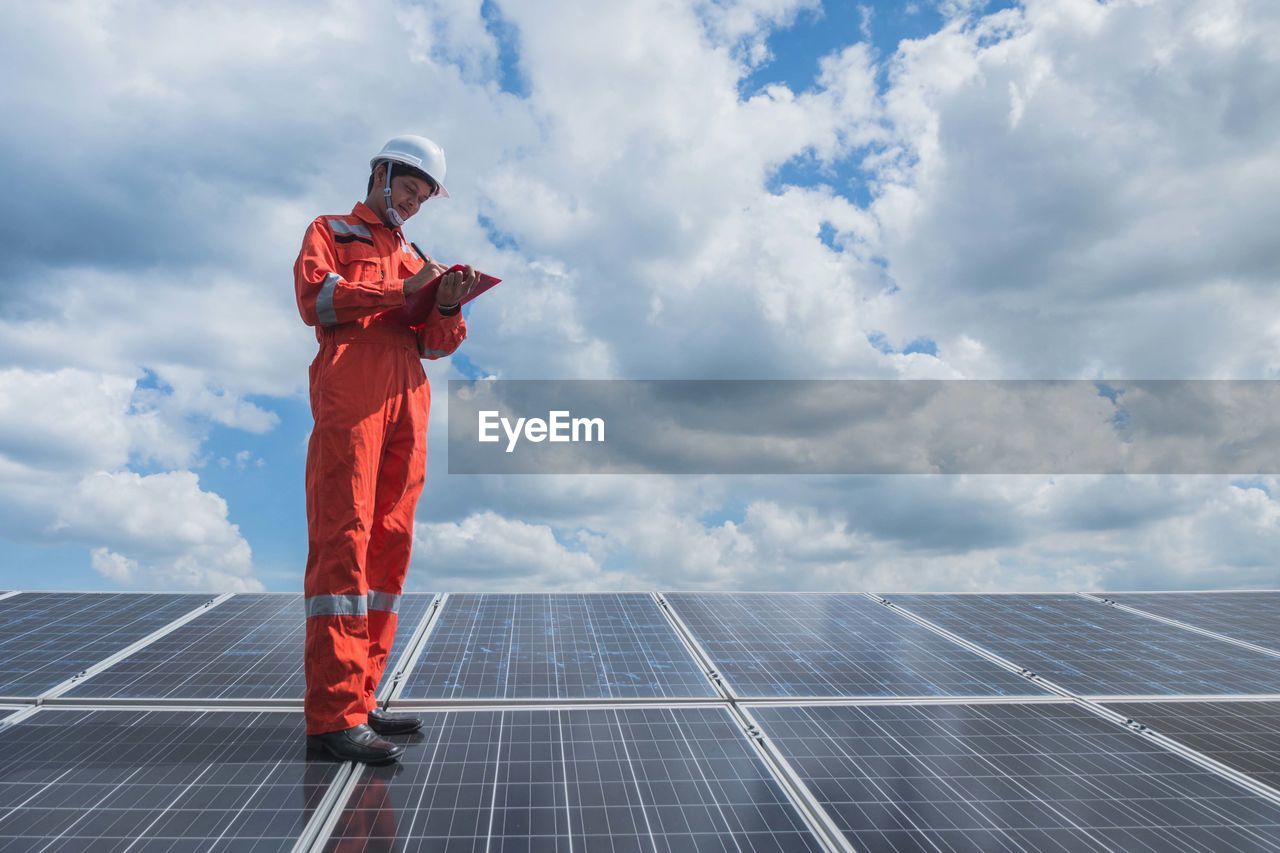 Low angle view of worker wearing reflective clothing while standing on solar panel against sky