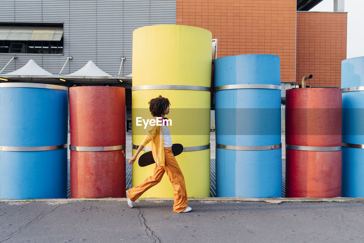 Young woman holding skateboard walking by multi colored pipes