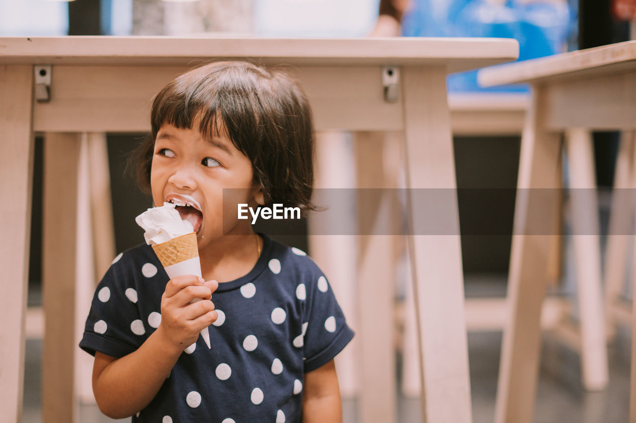 Cute girl eating ice cream while standing against tables