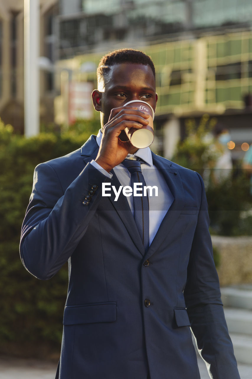 Serious adult formally dressed african american male manager drinking takeaway coffee while standing against blurred urban background of modern city