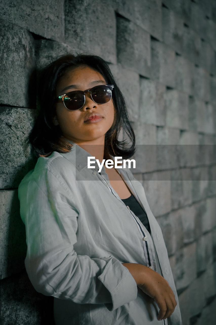 Portrait of young woman wearing sunglasses standing against wall