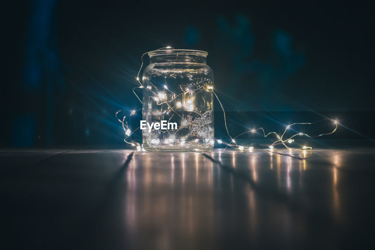 CLOSE-UP OF ILLUMINATED GLASS JAR ON TABLE AT NIGHT