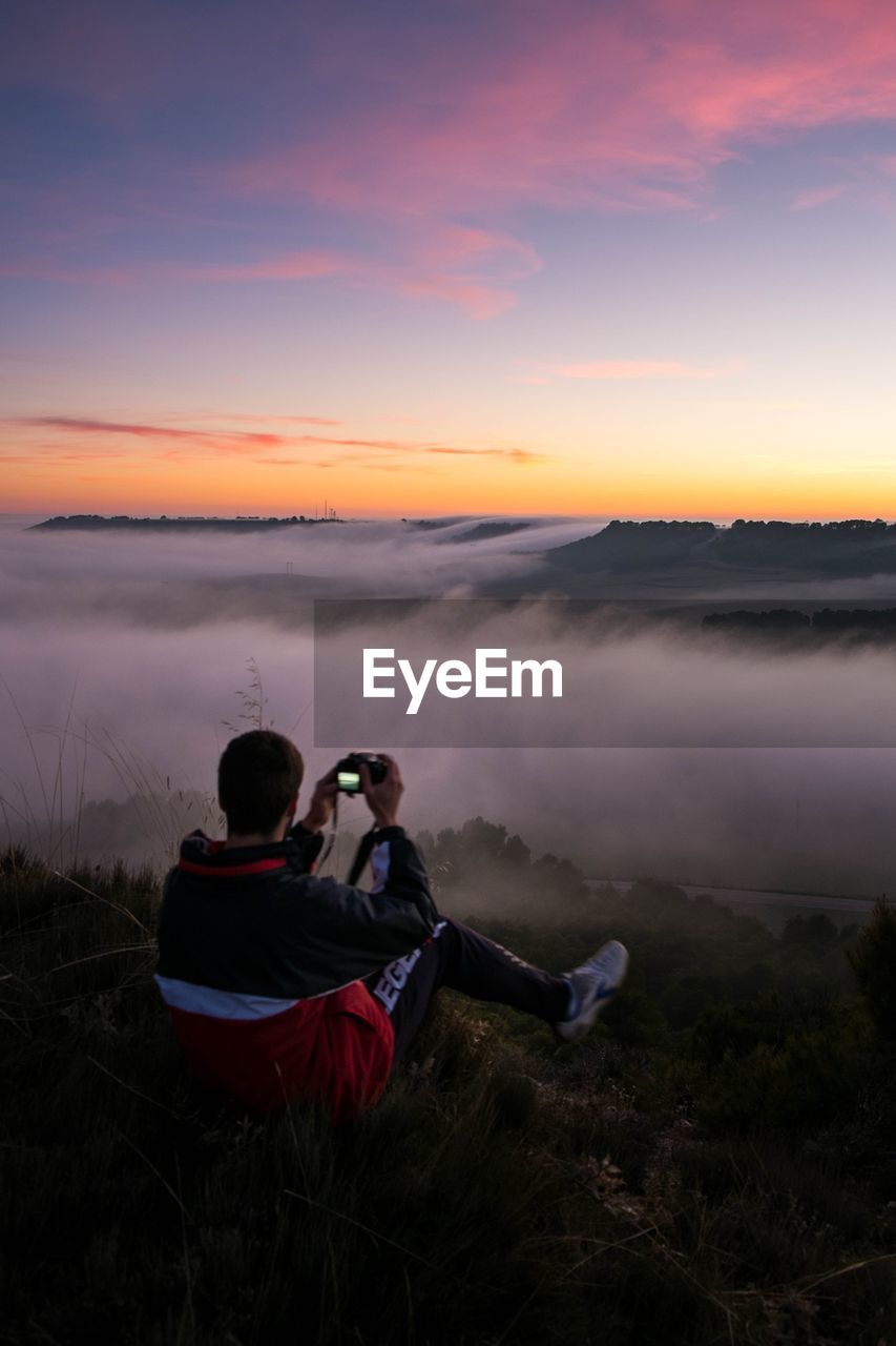 Rear view of man sitting on mountain against sky during sunset