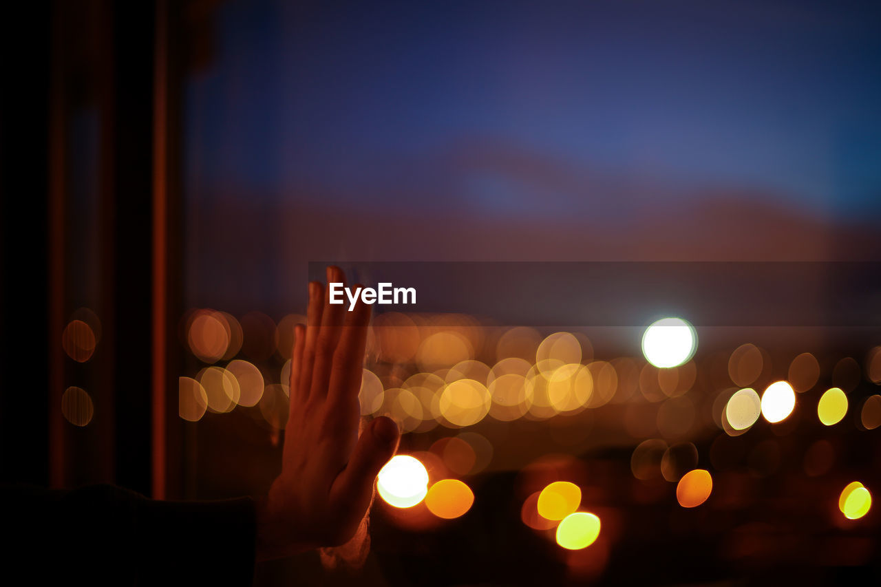 Cropped hand touching glass window against defocused light at dusk