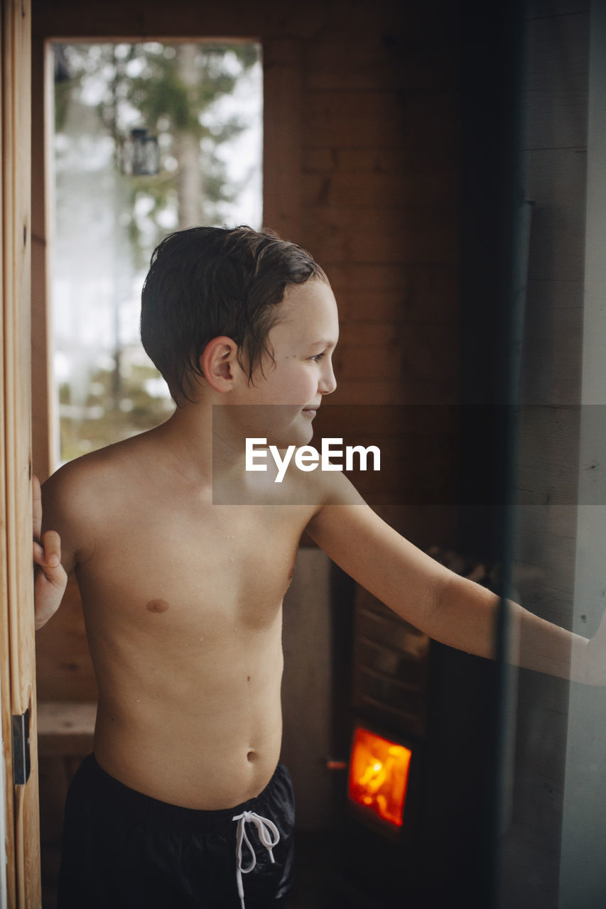 Smiling shirtless boy standing at doorway of sauna
