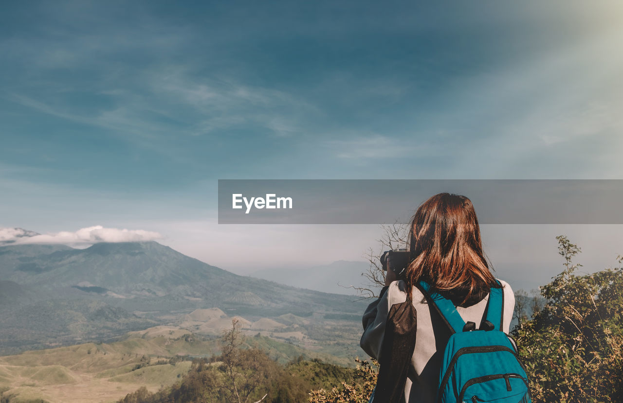 Rear view of woman with backpack photographing mountains while standing against sky