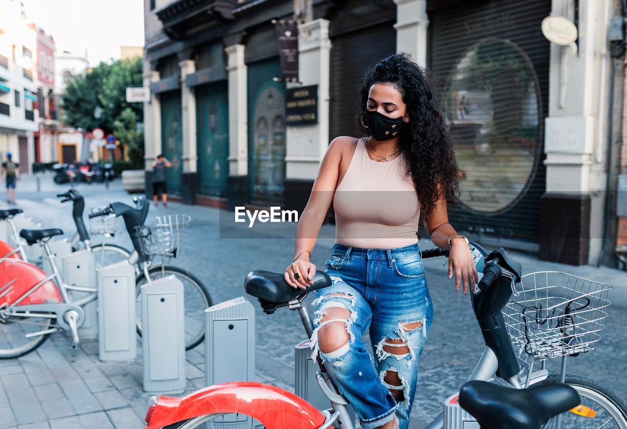 Side view of charming young latin female in jeans and protective mask standing near bicycle on city street and looking away