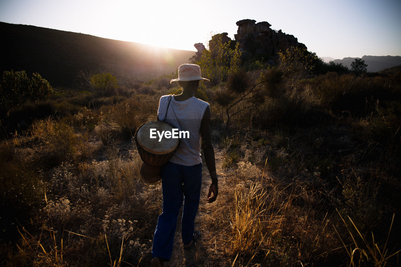 Rear view of man with drum walking on field during sunset