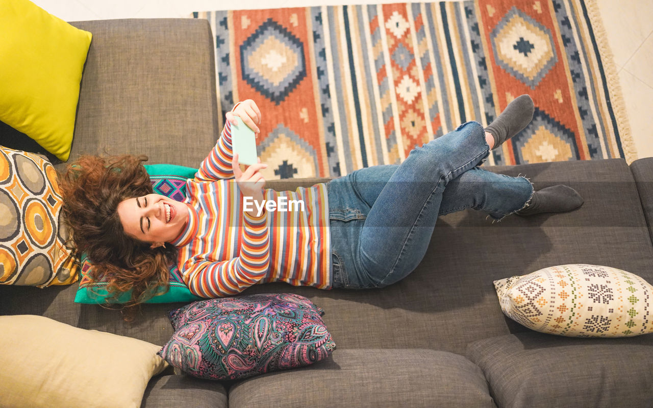 High angle view of cheerful woman taking selfie while lying on sofa at home
