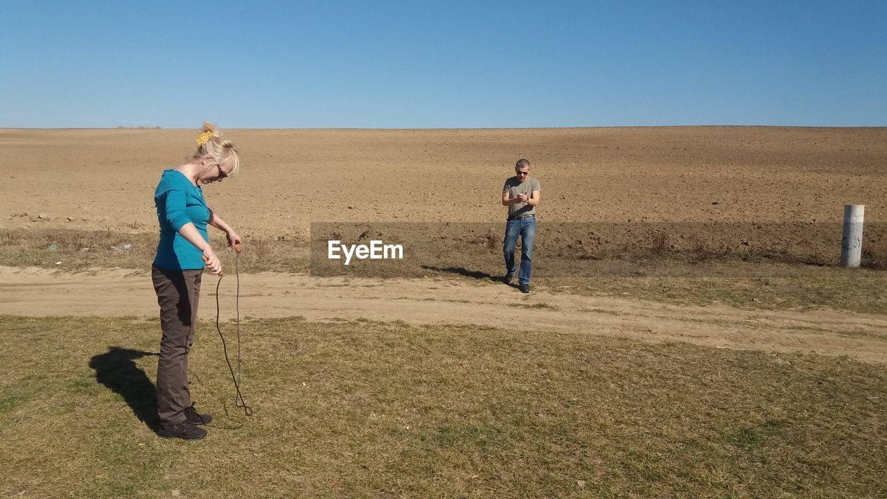 Woman looking at jumping rope while man walking on grassy field against clear sky