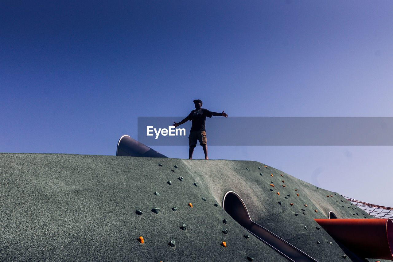 Low angle view of man standing on outdoor play equipment against clear blue sky