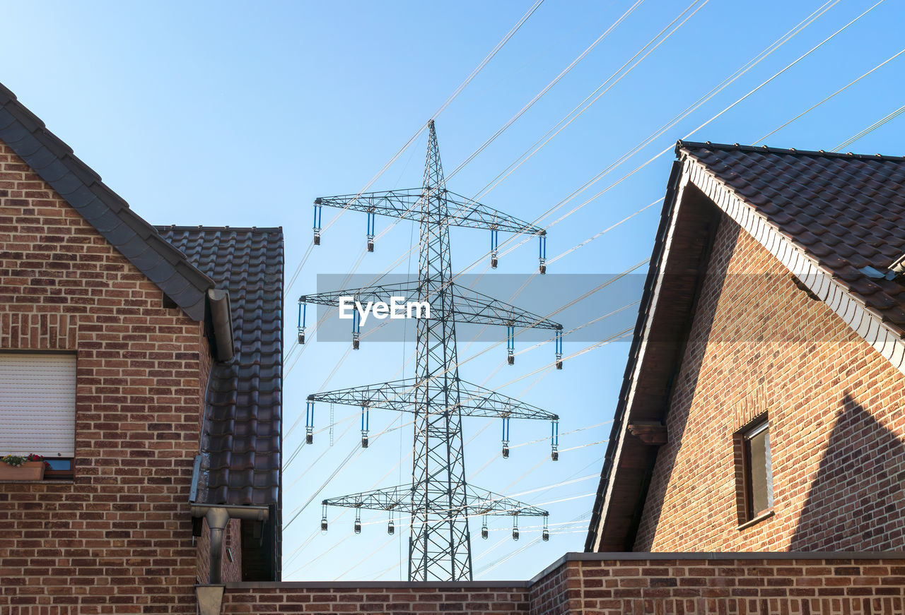 High voltage power pylon between houses - low angle view of building against sky