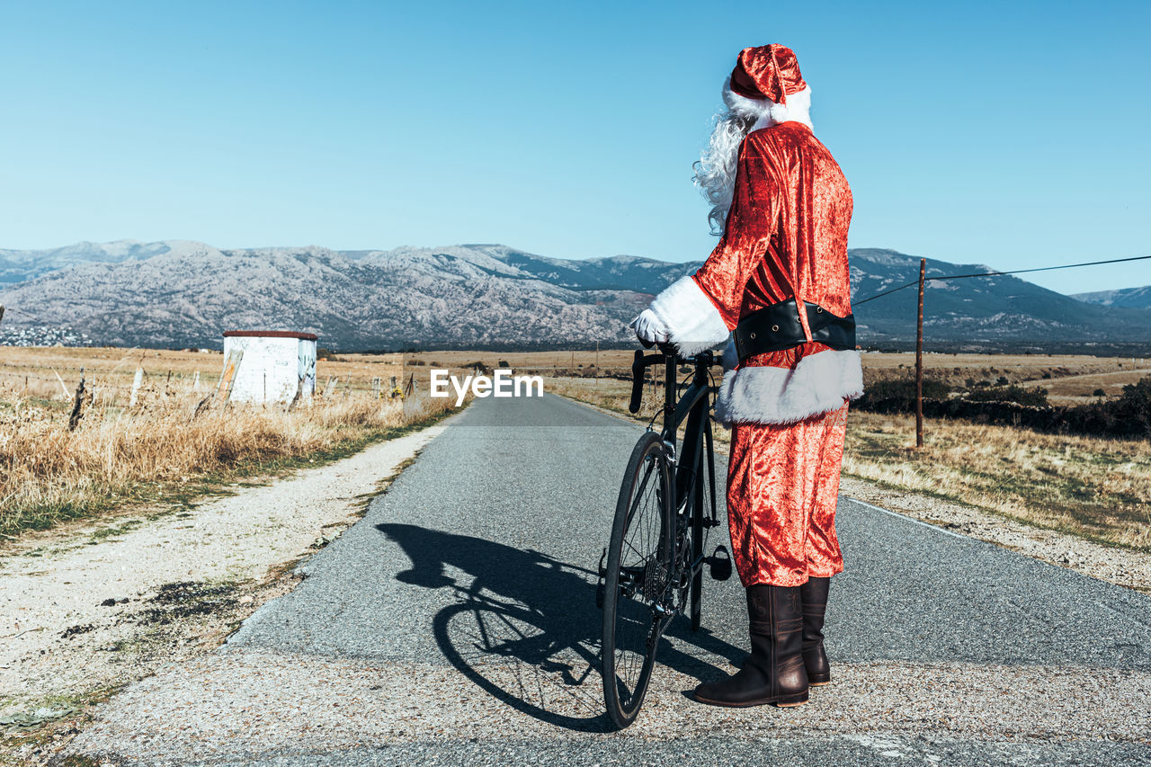 Back view of unrecognizable santa claus in red clothes standing with modern bicycle on empty road and looking away