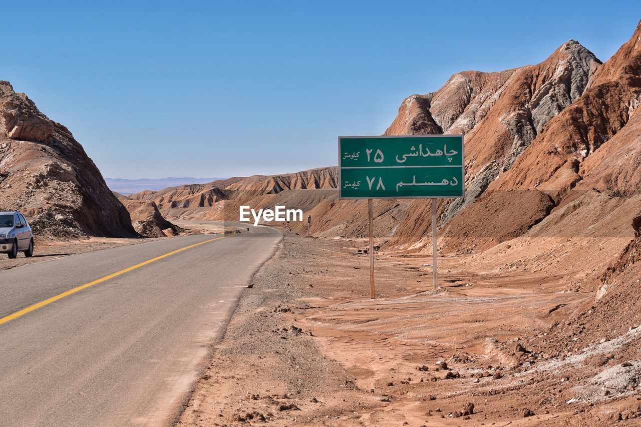 road, sign, transportation, sky, landscape, nature, desert, communication, wadi, environment, text, road sign, road trip, travel, natural environment, scenics - nature, clear sky, mountain, rock, travel destinations, valley, no people, symbol, land, geology, non-urban scene, blue, western script, soil, rock formation, day, mode of transportation, sunny, highway, guidance, outdoors, arid climate, climate, motor vehicle, information sign, car, beauty in nature, infrastructure, vehicle, arrow symbol, plateau, tourism, the way forward, sunlight