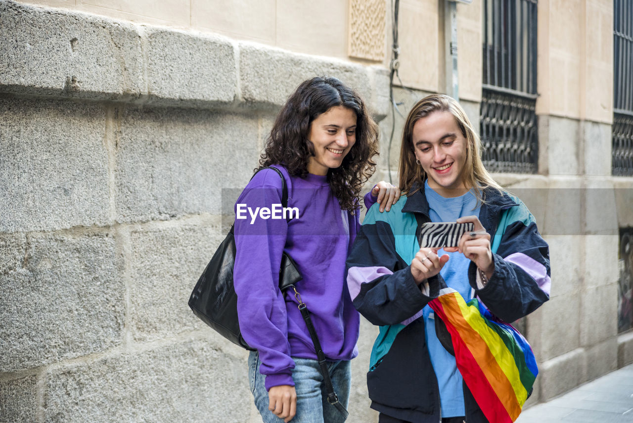 Two friends taking a selfie while walking in the street outdoors with lgbt flag.