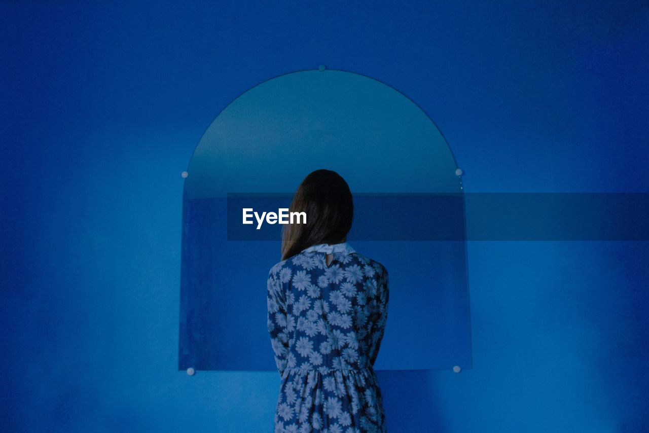 Rear view of woman standing against mirror on blue wall