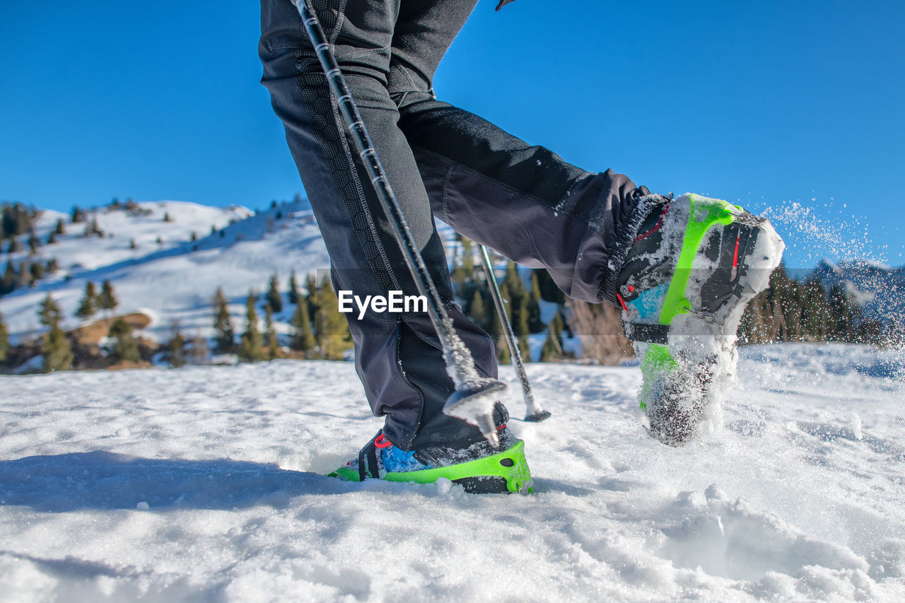 Low section of man walking on snow covered land against clear blue sky