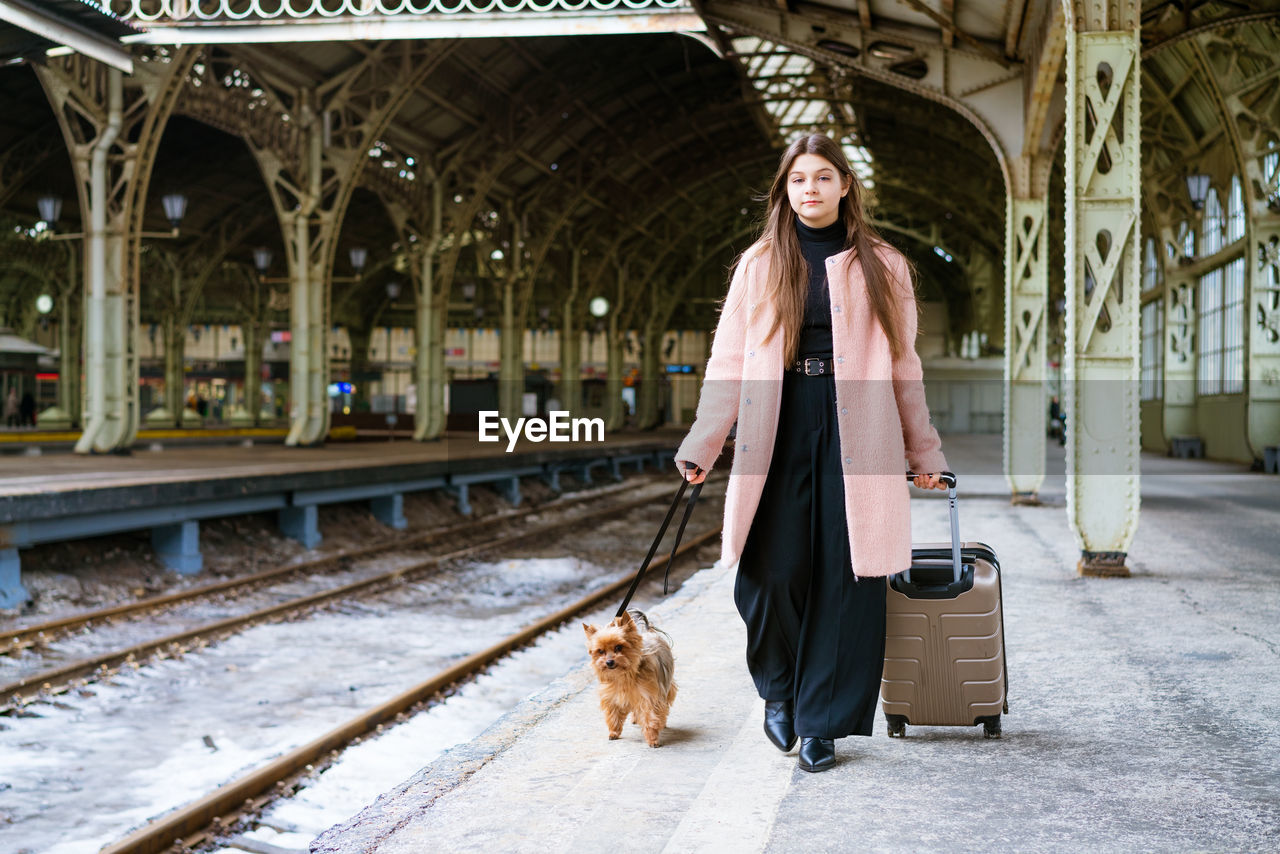 Beautiful young casual tourist woman with dog and suitcase waiting for train person