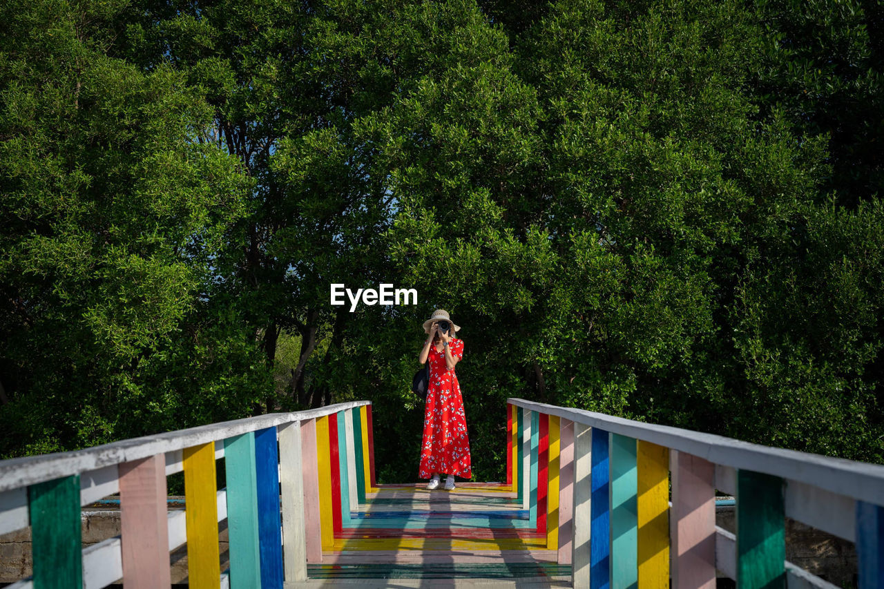 Woman photographing while standing on footpath against trees