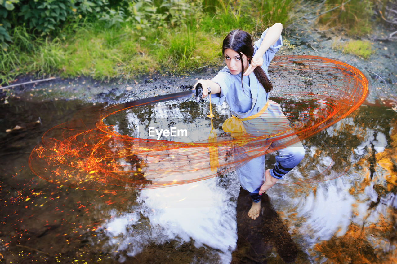 Young woman practicing kung fu with sword while standing on puddle in forest
