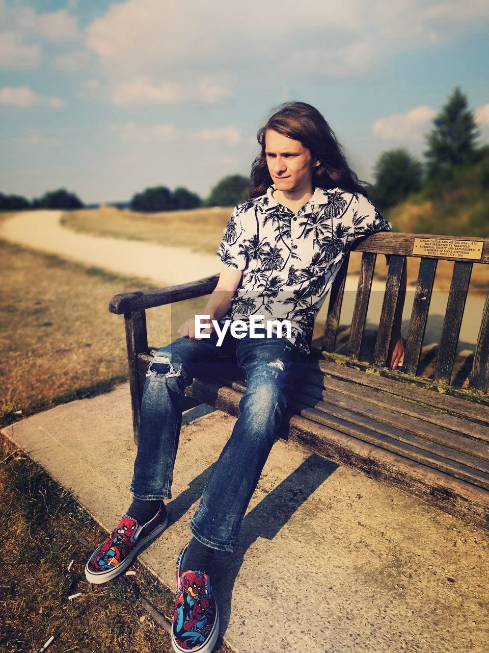 FULL LENGTH OF YOUNG WOMAN SITTING ON BENCH AT FIELD