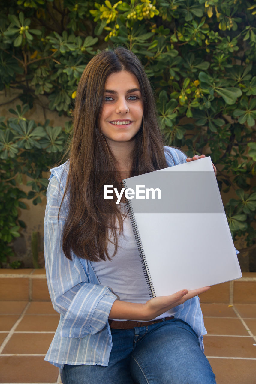 Portrait of smiling woman holding spiral notebook against plants