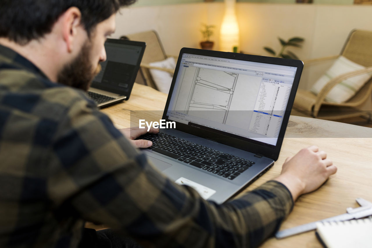 Male carpenter making drawing on laptop while sitting at desk in industry