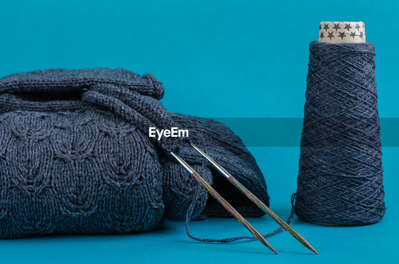 Cone of gray wool yarn and part of knitted sweater on knitting needles, turquoise background