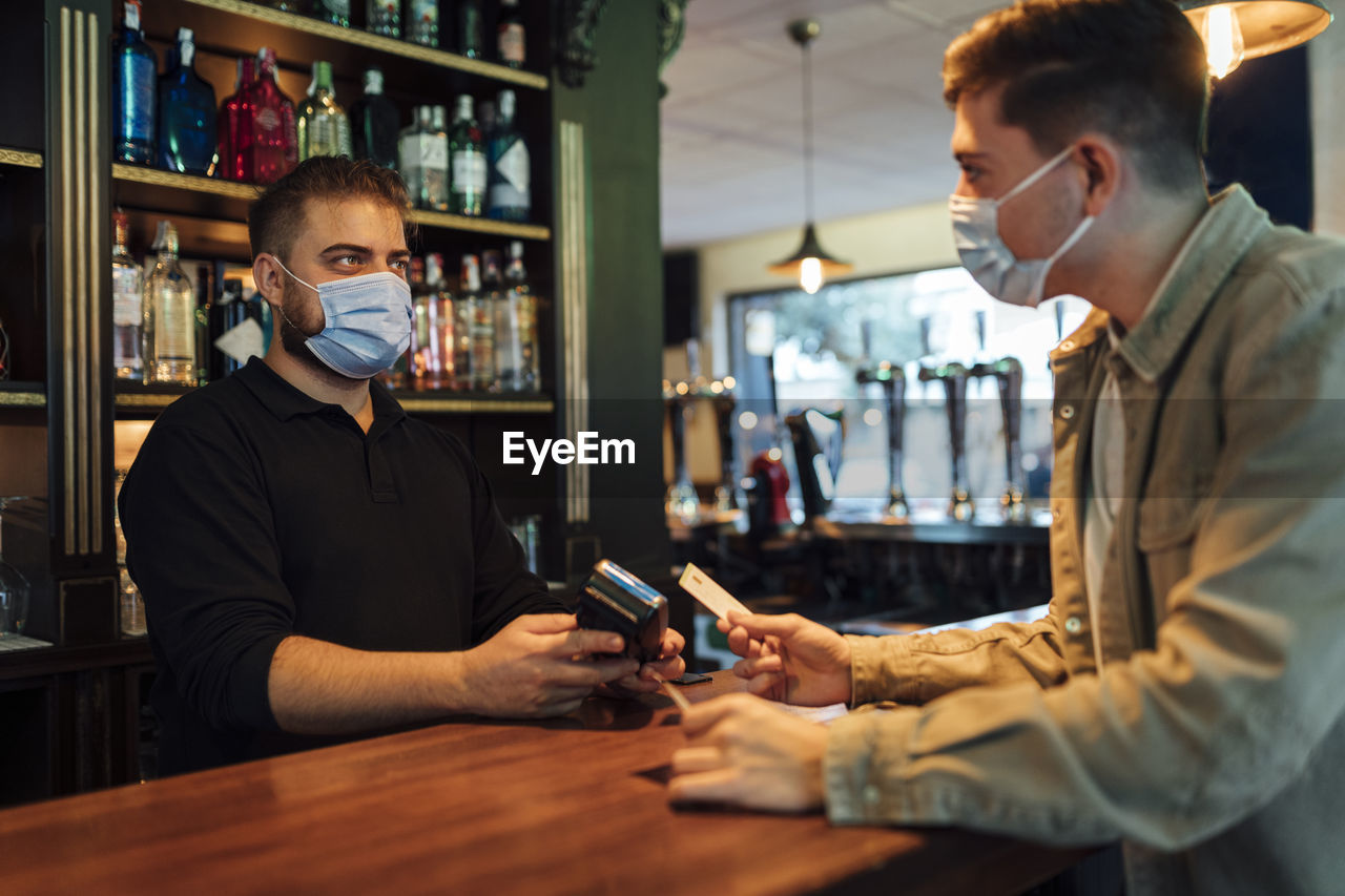 Male customer paying with credit card to waiter in bar during pandemic