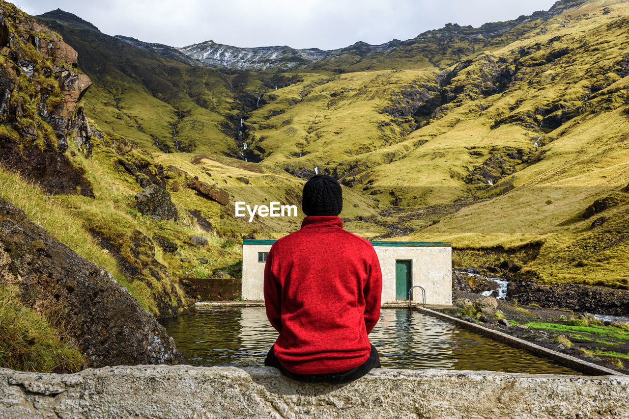 REAR VIEW OF MAN SITTING ON MOUNTAIN LOOKING AT VIEW