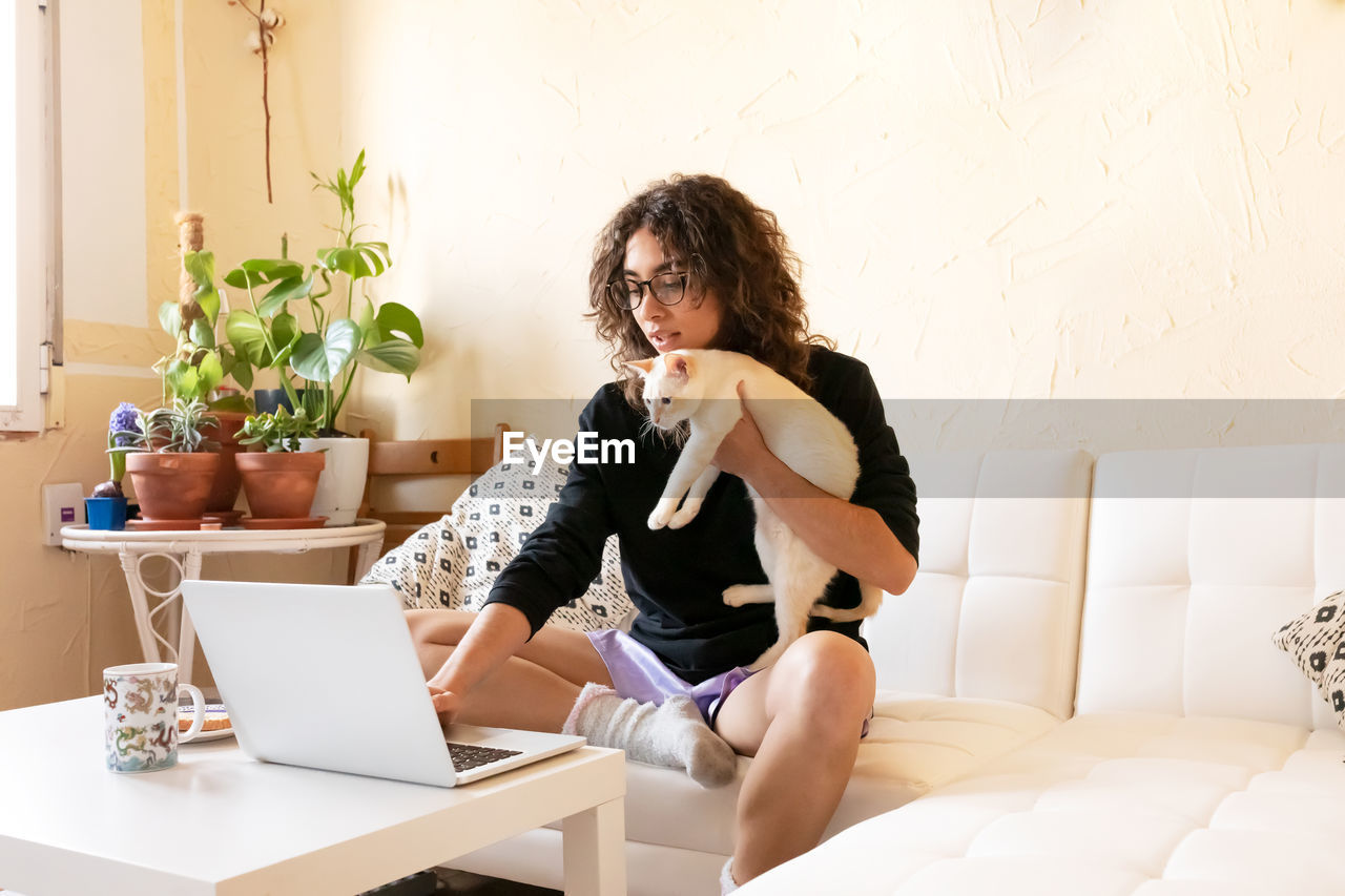 Young ethnic female remote worker in homey outfit and glasses holding cute cat while using laptop in living room