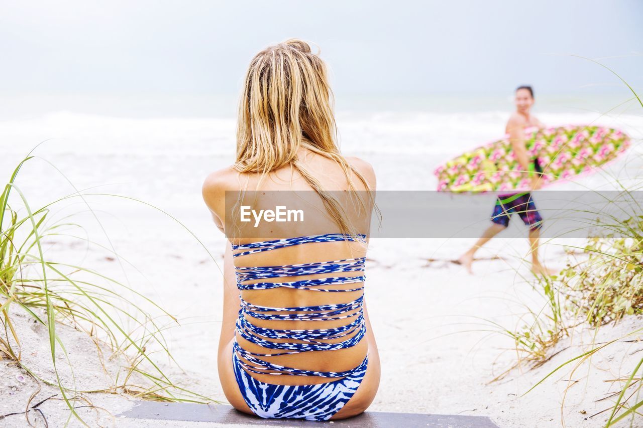 Rear view of woman sitting in front of man with surfboard walking at beach