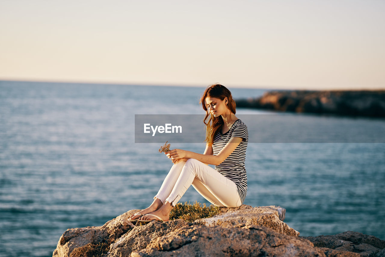 WOMAN SITTING ON ROCK AGAINST SEA